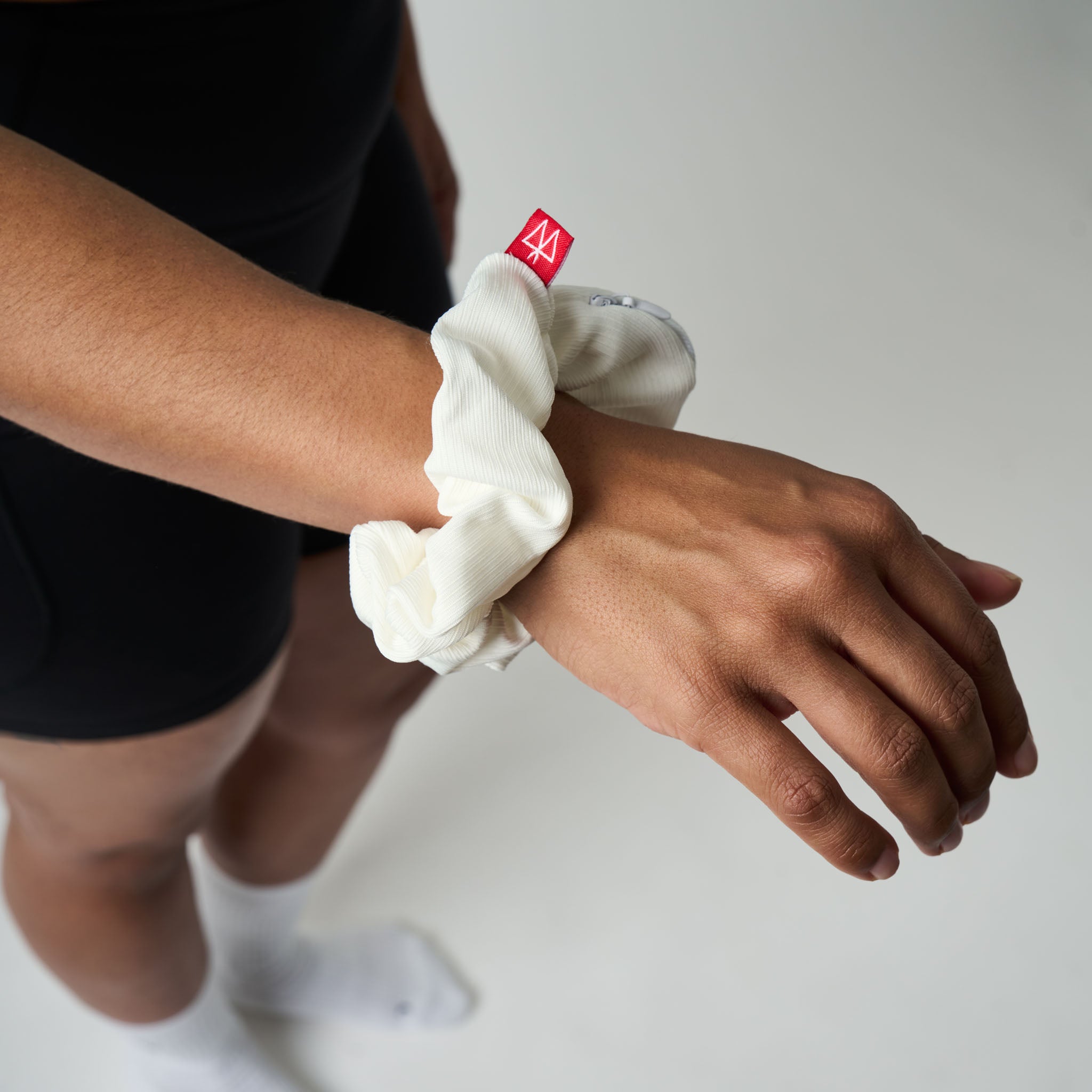 Person wearing a white scrunchie on their wrist with a red logo, against a neutral background.