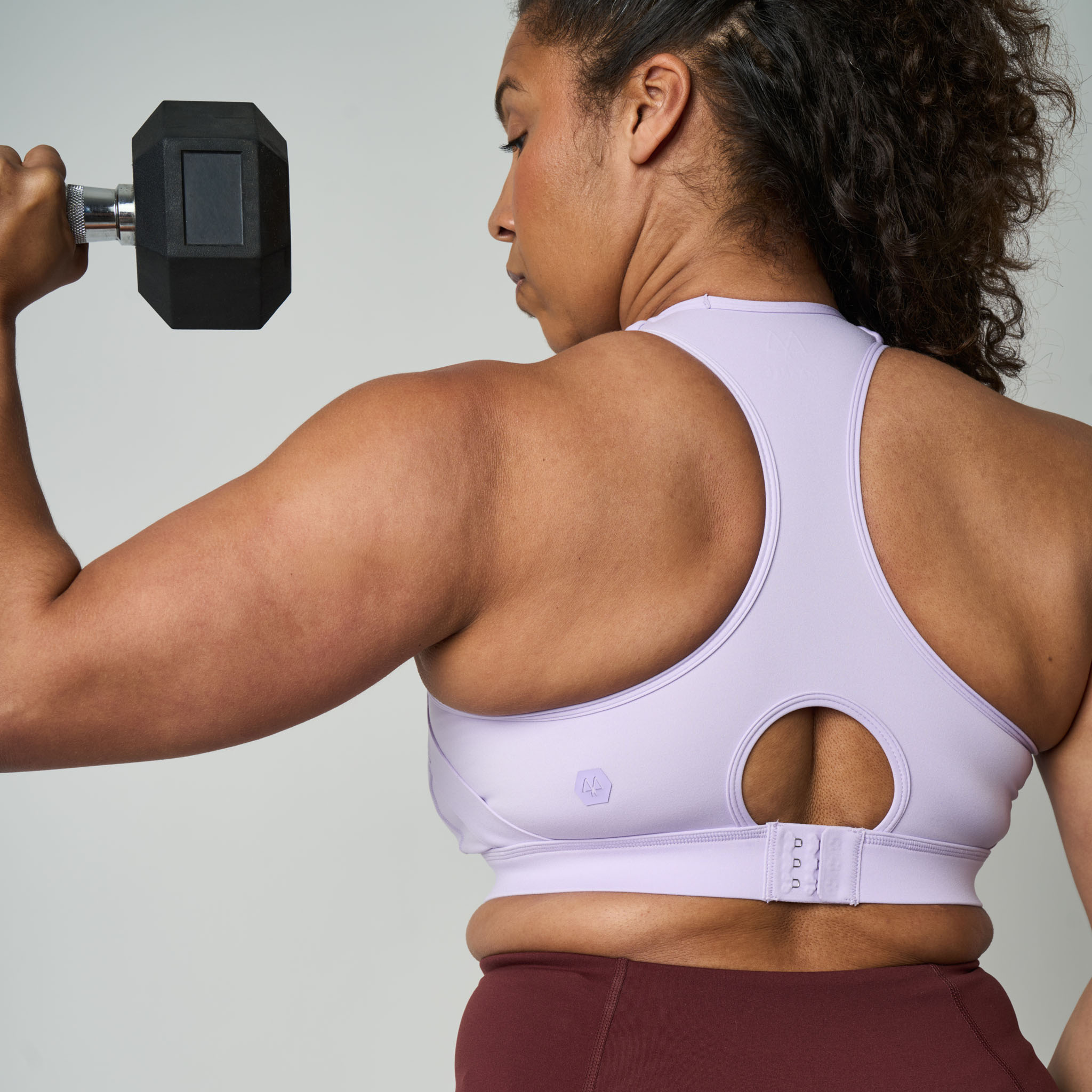 Woman lifting a dumbbell wearing a white sports bra and maroon leggings on a light gray background